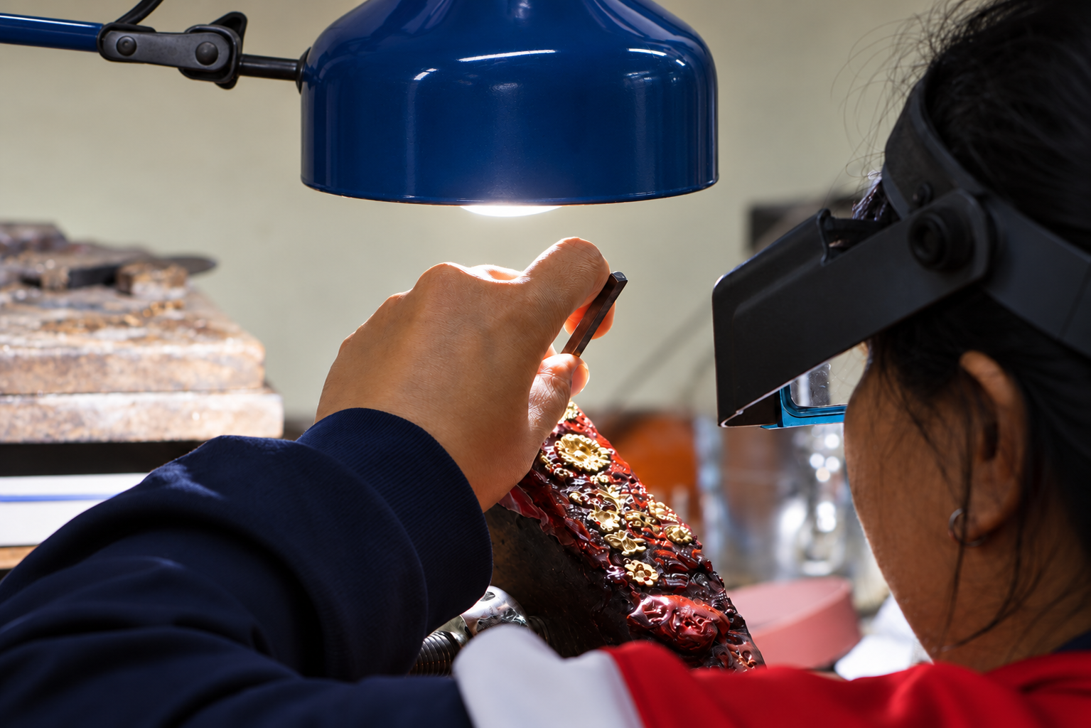 Skilled jewelry artisan wearing a magnifying visor carefully working on intricate gold-colored metal details under a workshop lamp during the jewelry hand-finishing process.