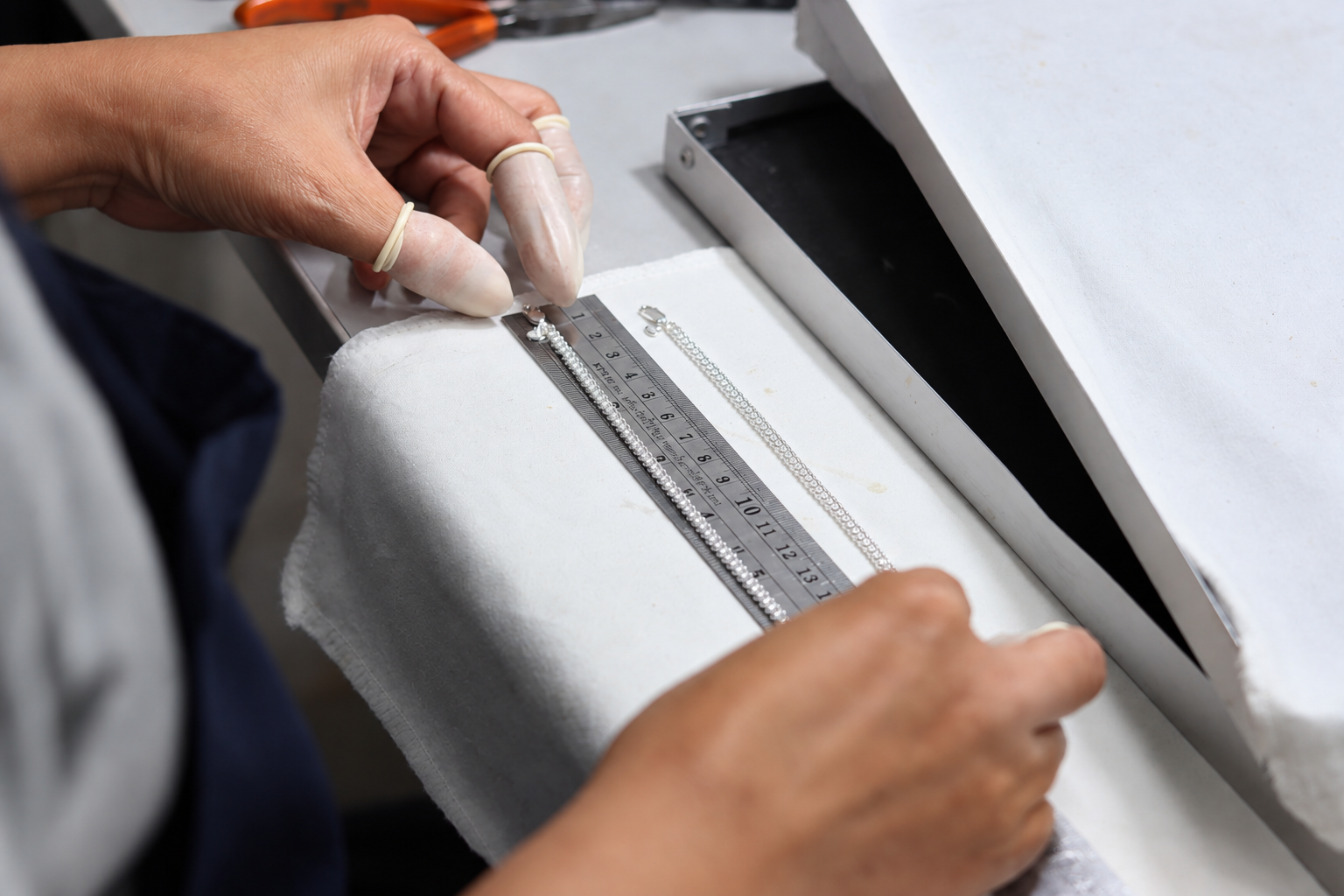 Jewelry quality control worker measuring a silver-colored chain against a metal ruler to check its length during jewelry production.