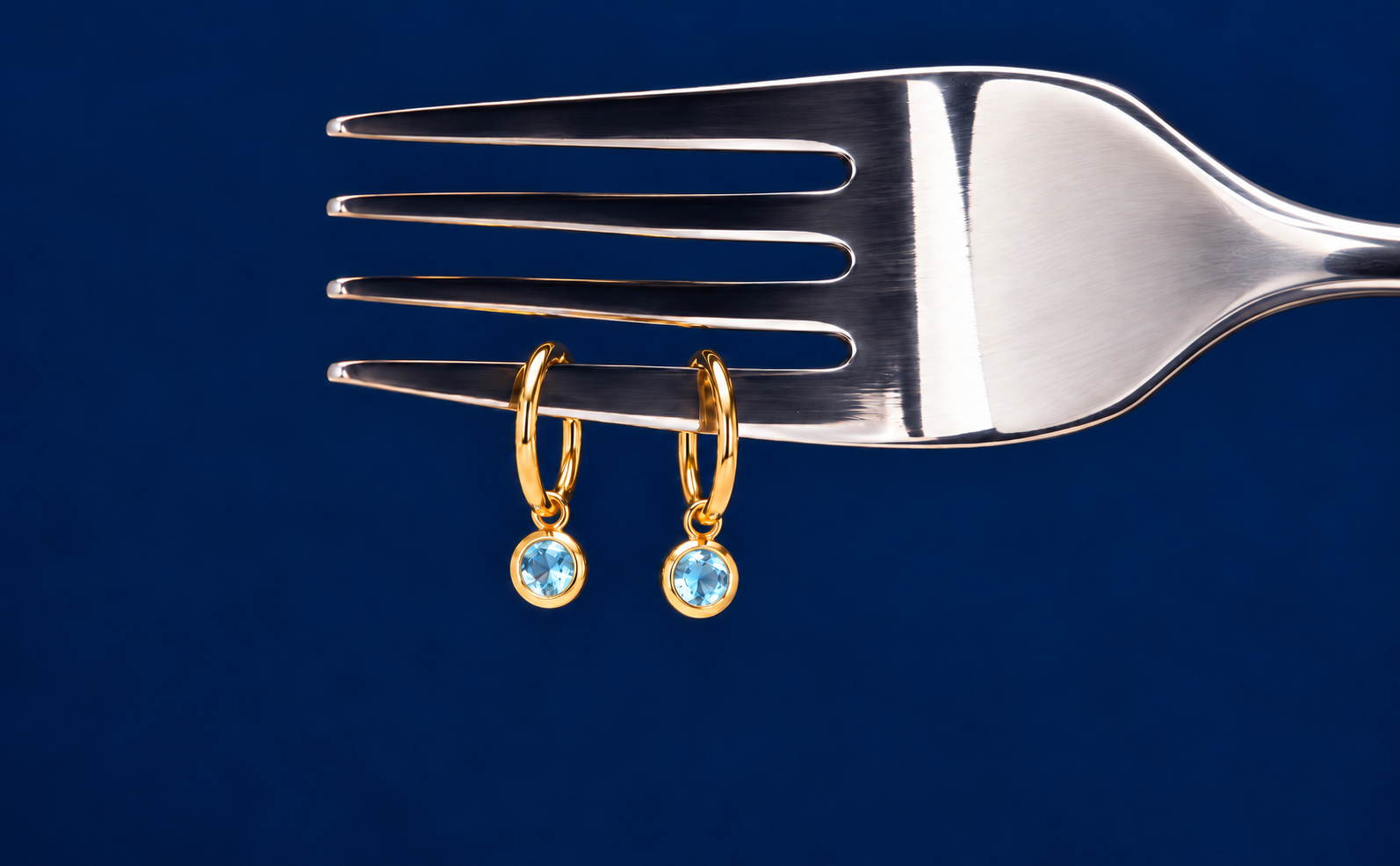 Gold hoop drop earrings with round soft aquamarine blue gemstones in gold bezel settings, displayed on a silver fork against a navy blue background.