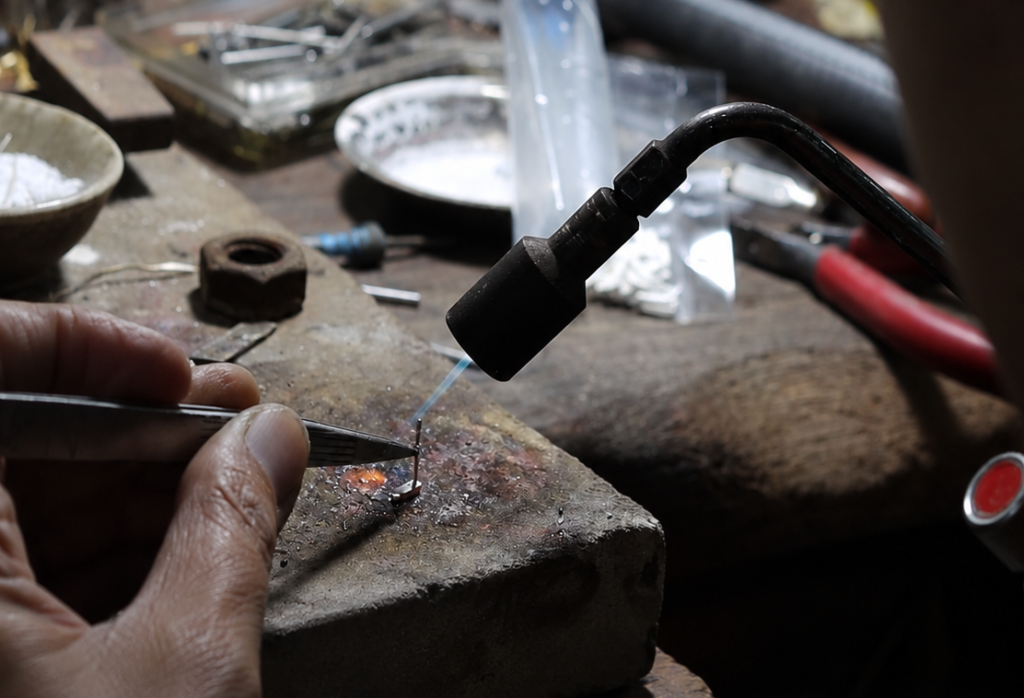 Jewelry artisan using a precision torch and tweezers to solder a small metal component during custom jewelry manufacturing.
