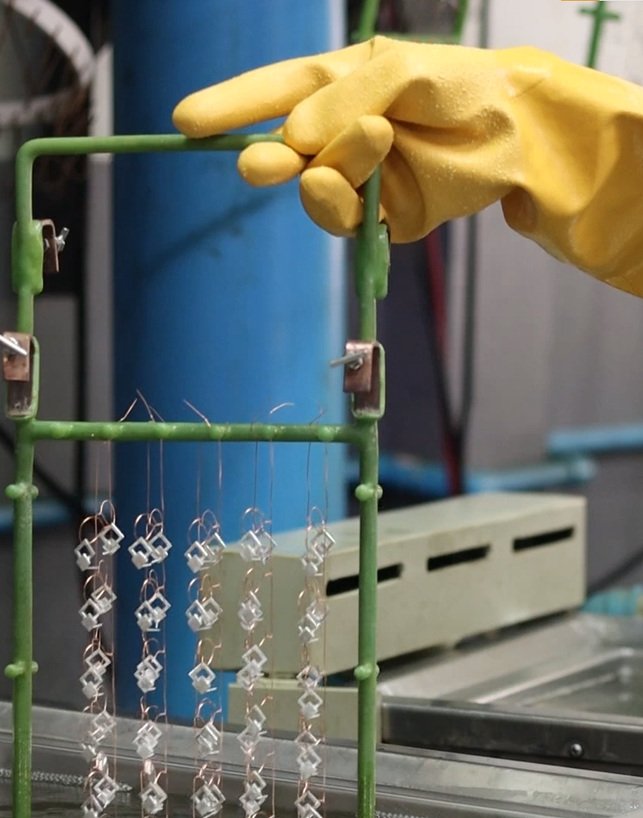 Jewelry manufacturing worker wearing yellow protective gloves holding metal jewelry components on a rack above a plating tank.