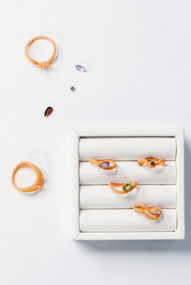 Custom jewelry ring models with colorful gemstones displayed on a white jewelry tray, alongside loose gemstones and ring settings during the jewelry design process.