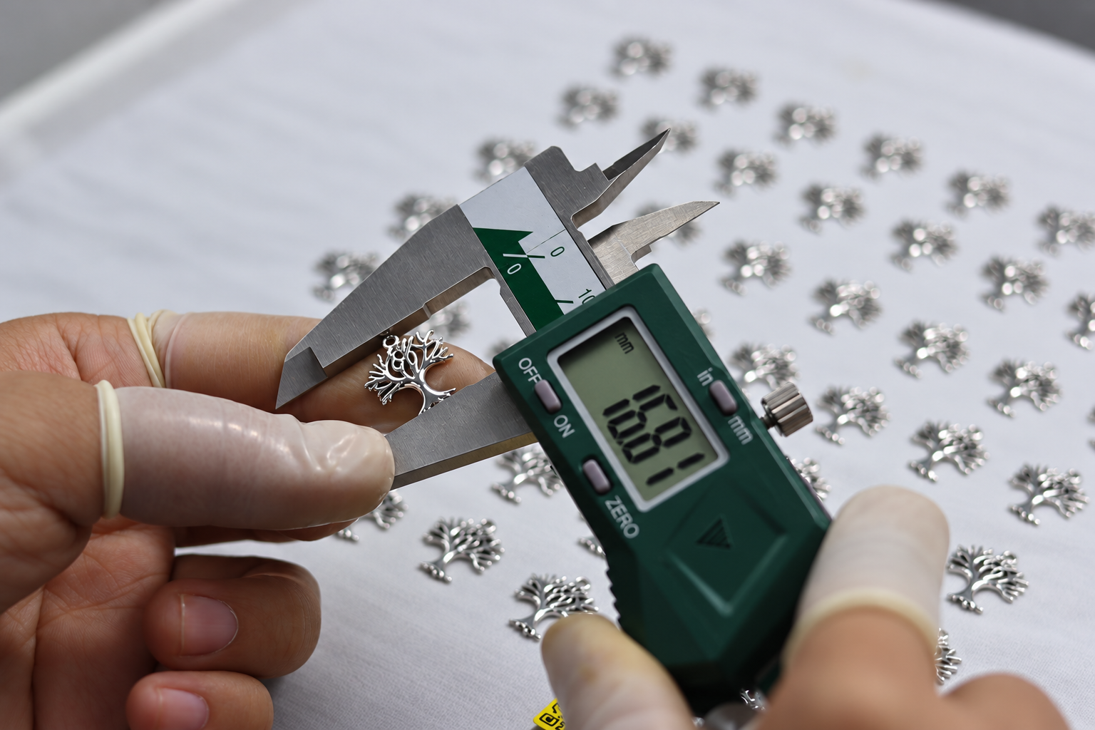 Jewelry quality control worker using a digital caliper to measure a tree-shaped metal jewelry component during dimensional inspection.