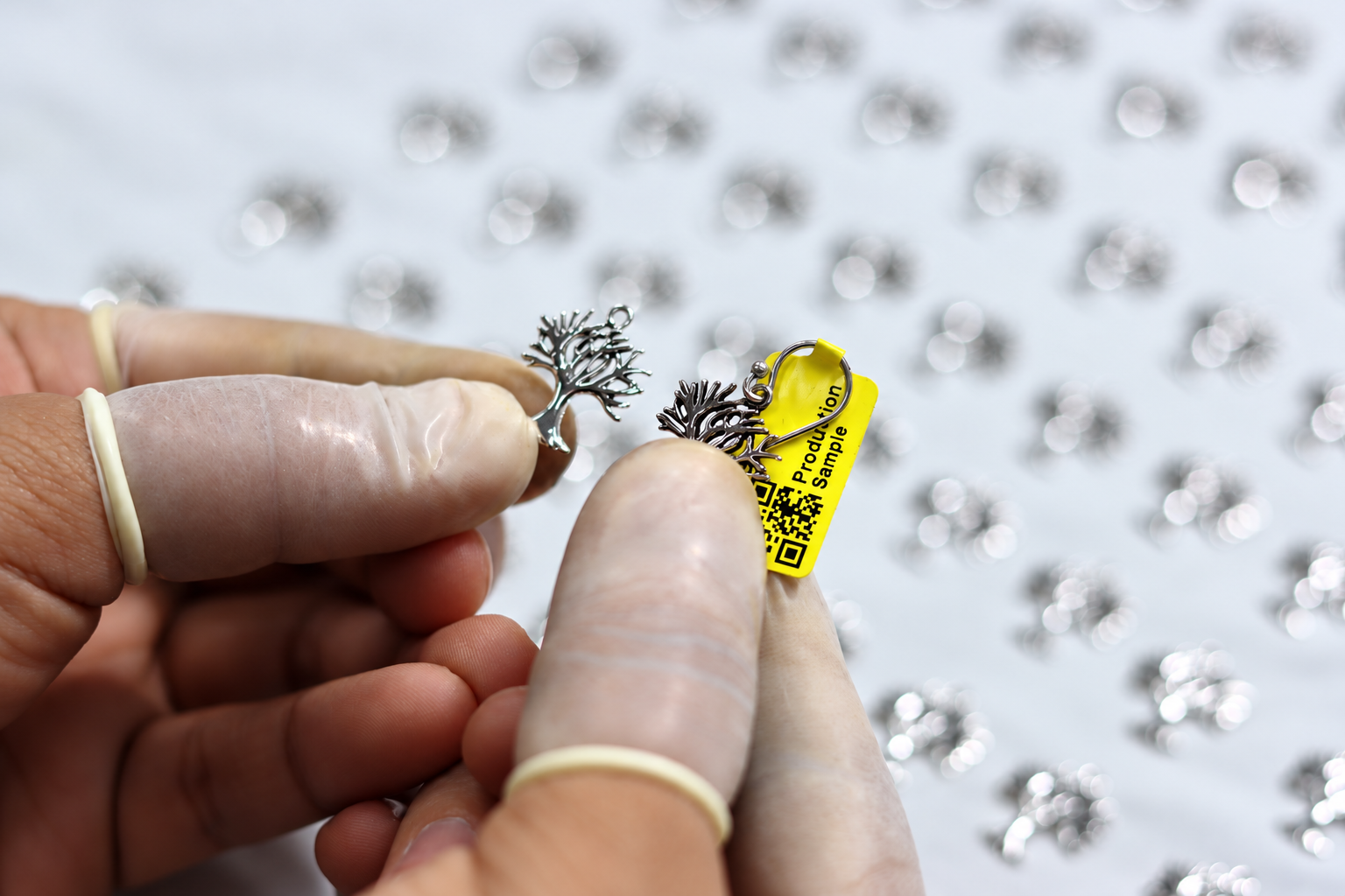 Jewelry quality control worker comparing two silver-colored tree-shaped jewelry charms, including a tagged production sample, during visual inspection.