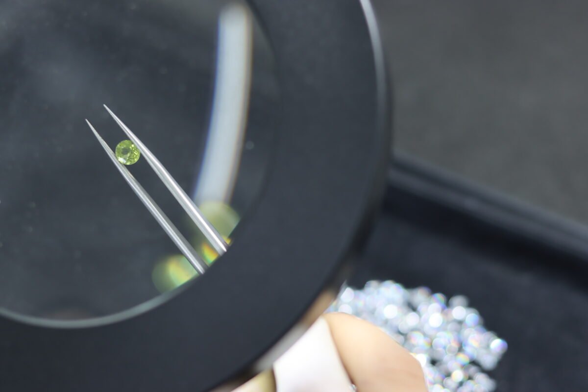 Close-up of a small green gemstone held with precision tweezers under a magnifying lens during a jewelry materials quality inspection.